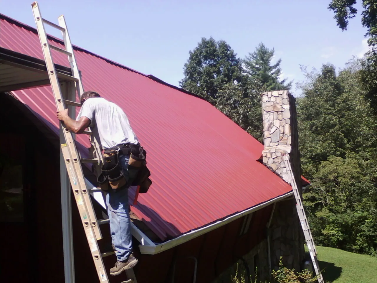 Roofer on a ladder beside a red metal roof and stone chimney — Soffit & Fascia Repair by Roofing Brothers in Burtonsville